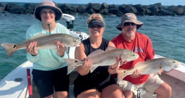 Three anglers each holding a redfish on a boat in front of the jetty rocks
