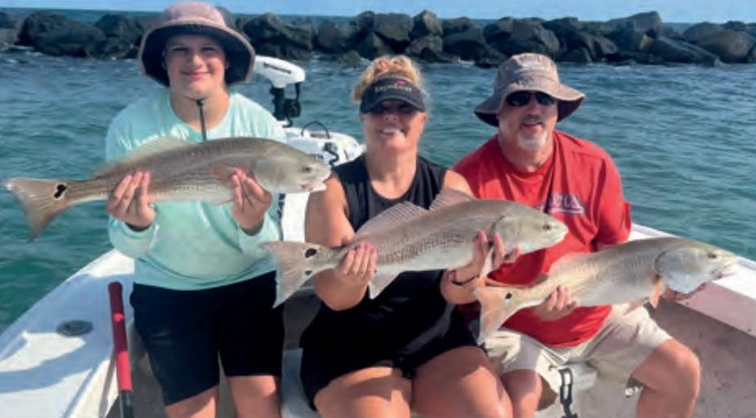 Three anglers each holding a redfish on a boat in front of the jetty rocks