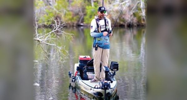 An angler standing on a fishing kayak on a tree-lined river