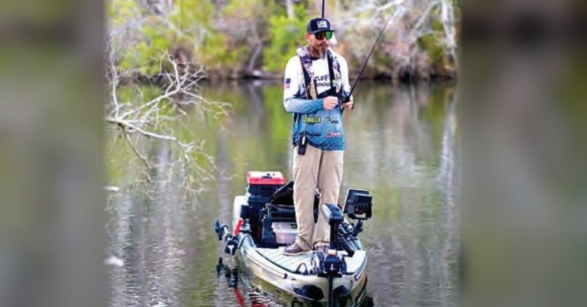 An angler standing on a fishing kayak on a tree-lined river