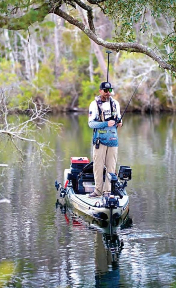 An angler standing on a fishing kayak on a tree-lined river